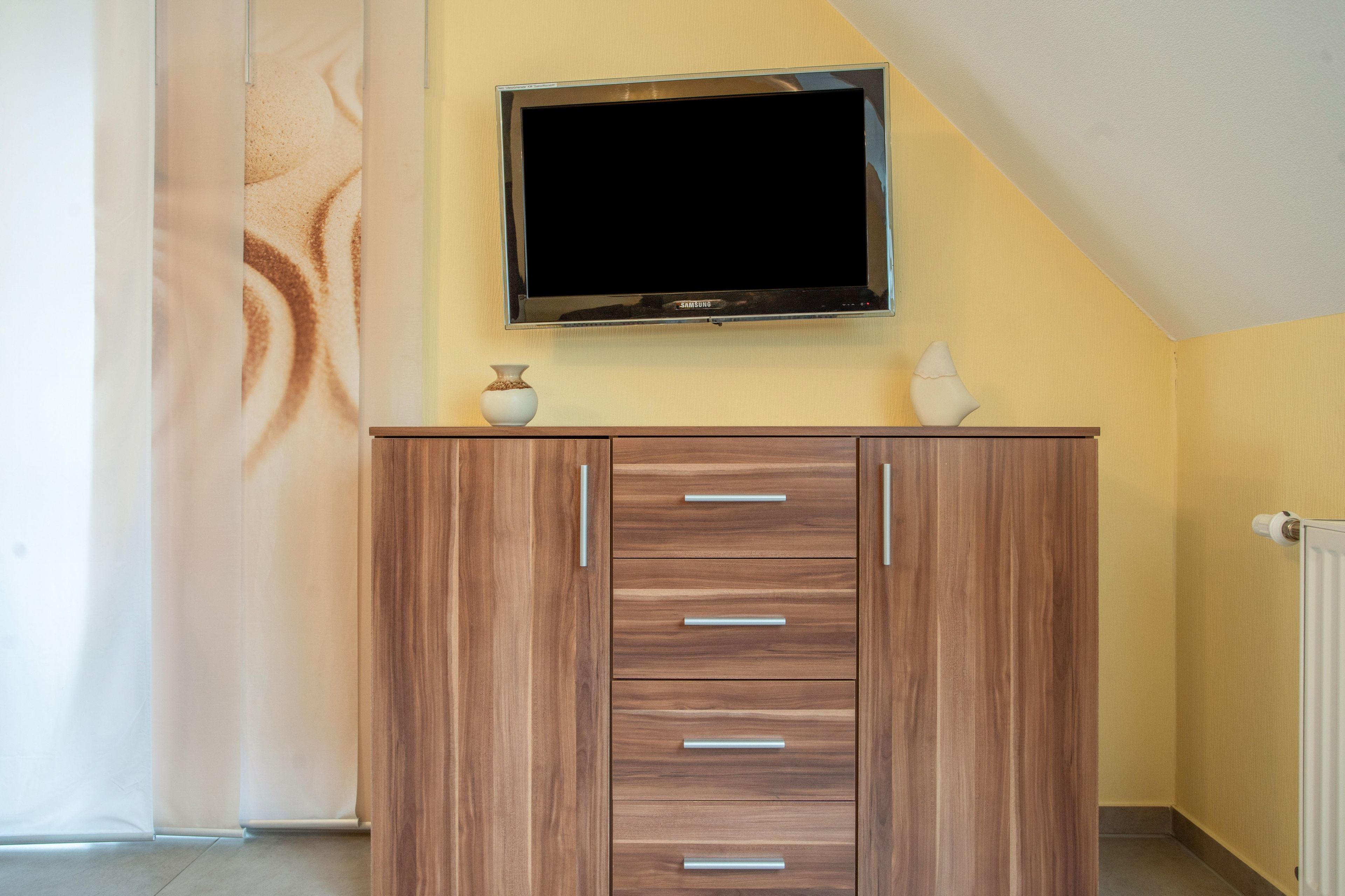 Een kamer met een aan de muur bevestigde televisie, een houten kast met lades en een radiator.