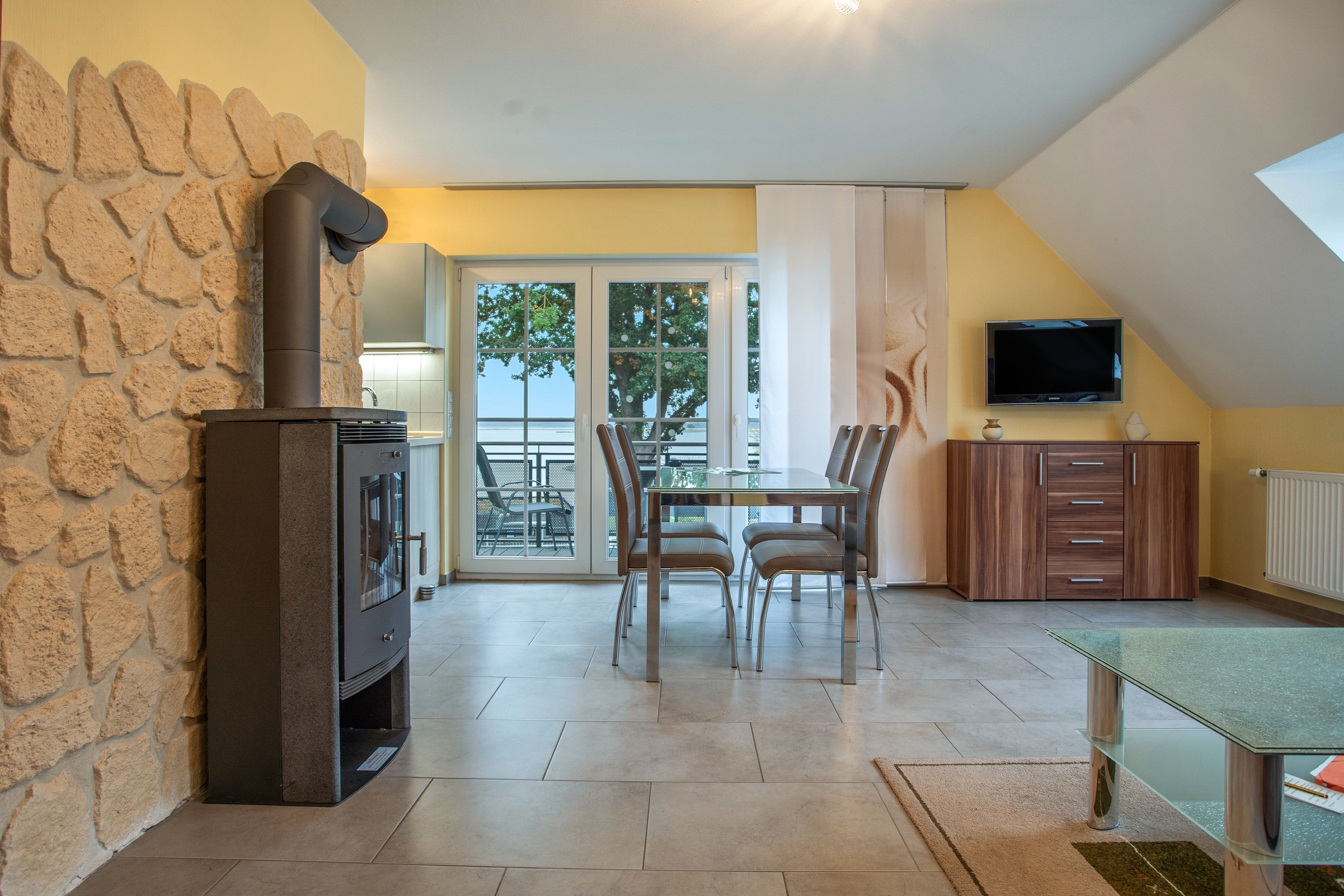 Un comedor con pared de piedra, chimenea, mesa con sillas, televisor y vistas al exterior.