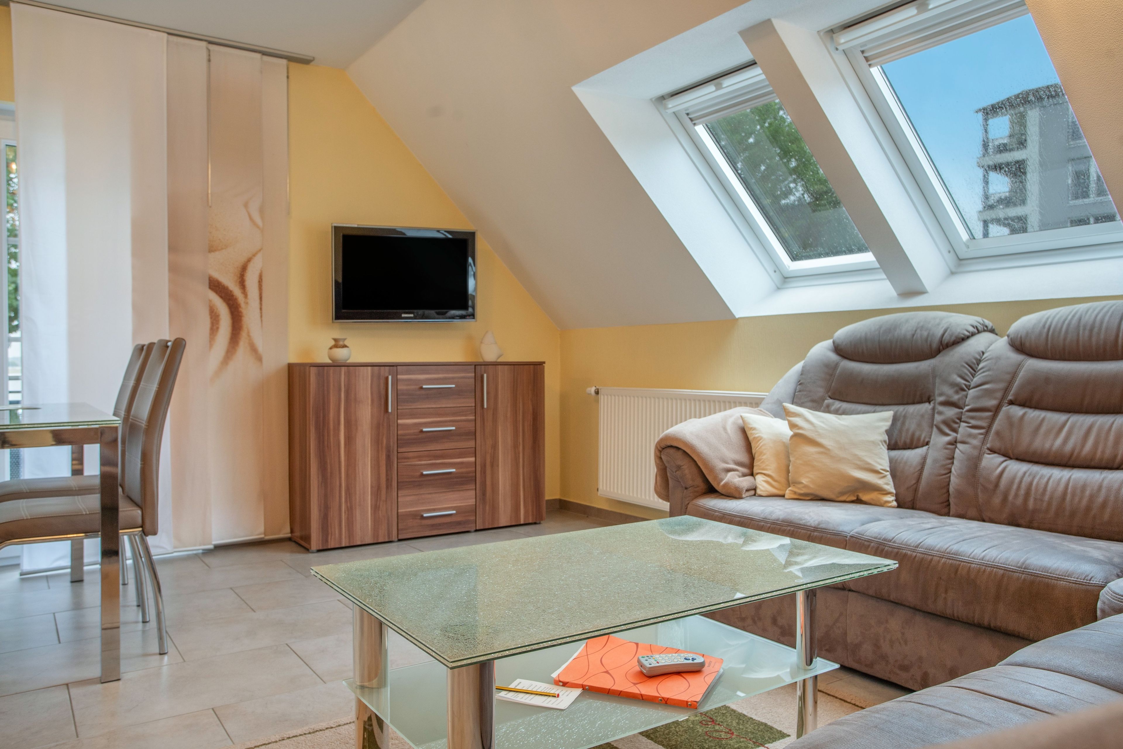 Una sala de estar moderna con una mesa de centro de vidrio, un sofá, un televisor montado en la pared y un ventanal con vistas al exterior.