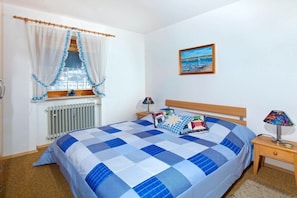 Ein Schlafzimmer mit einem großen Bett, einem Fenster mit Vorhängen, einem Heizkörper, einem Nachttisch mit einer Lampe und einem Gemälde an der Wand.