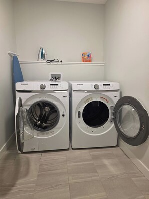 Two white Samsung front-loading washing machines in a laundry room with a blue ironing board and a shelf with laundry supplies. - Spacious Family House in Edmonton - Perfect for a Comfortable Stay (Edmonton)