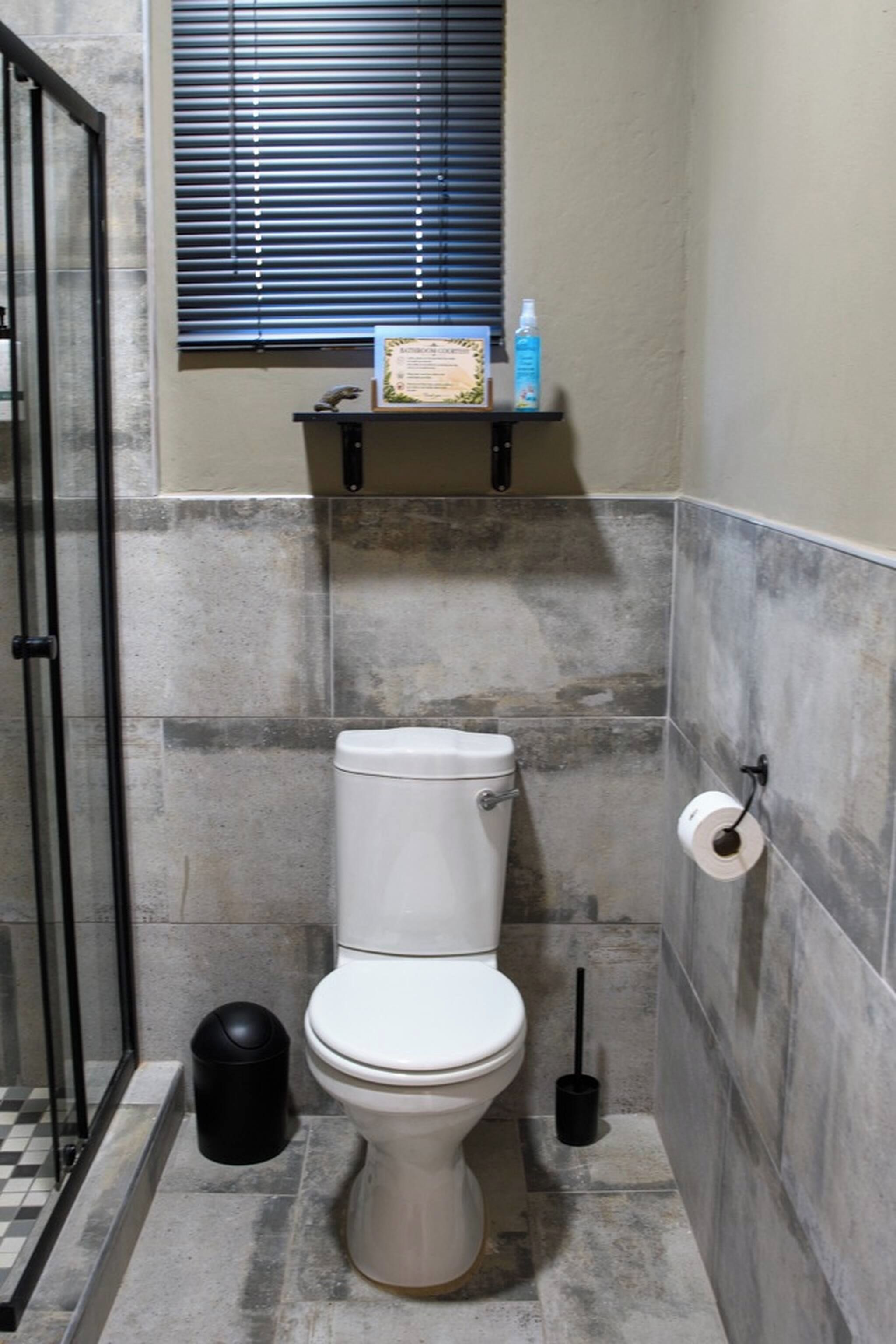 A bathroom with a toilet, a trash bin, a toilet paper holder, and a window with blinds.