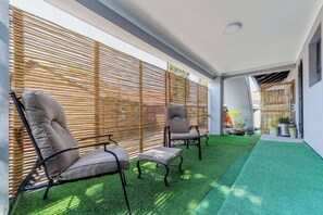 Zona d'estar a l'aire lliure amb dues cadires i una taula petita, envoltada de persianes de bambú i gespa artificial.