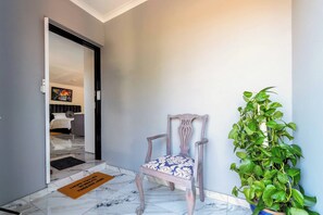 Um corredor com uma porta dando para um quarto, uma cadeira com almofada estampada, uma planta em vaso e um tapete de entrada com texto.