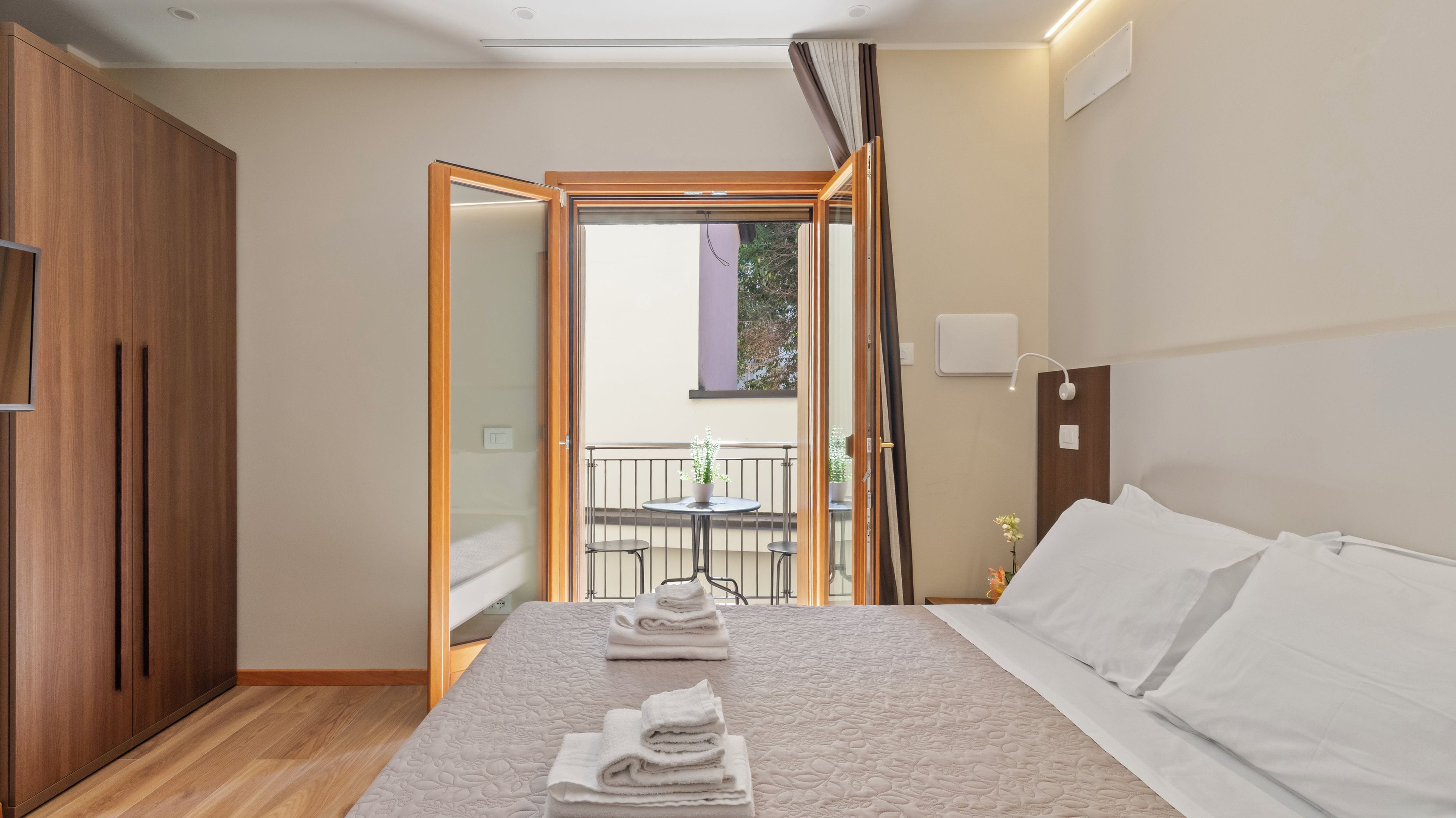 A hotel room with a bed, a wooden wardrobe, a balcony with a table and chairs, and a potted plant.