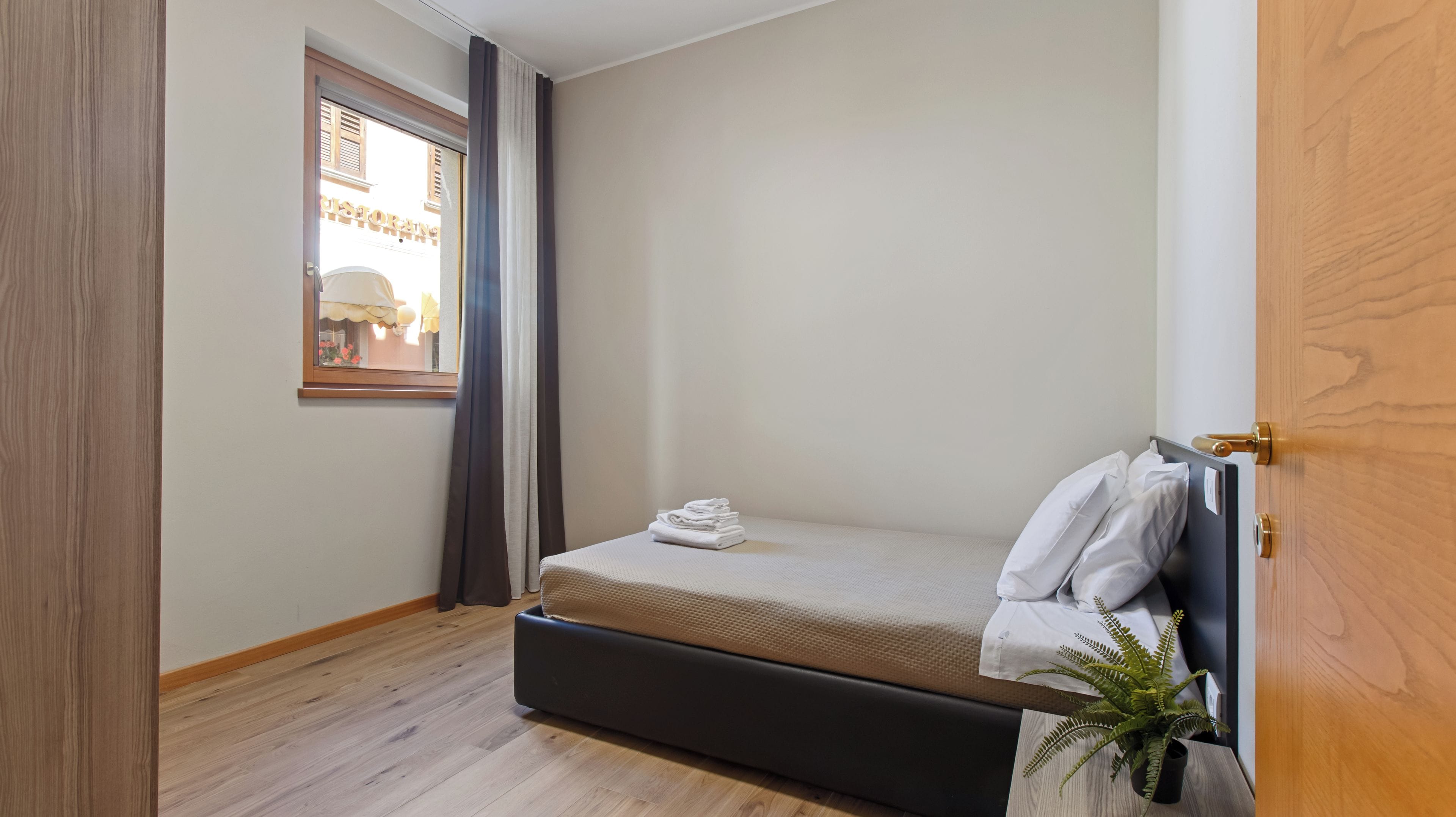 Una habitación pequeña con cama individual, suelo de madera, ventana con cortinas, planta en la mesita de noche y puerta con pomo de latón.