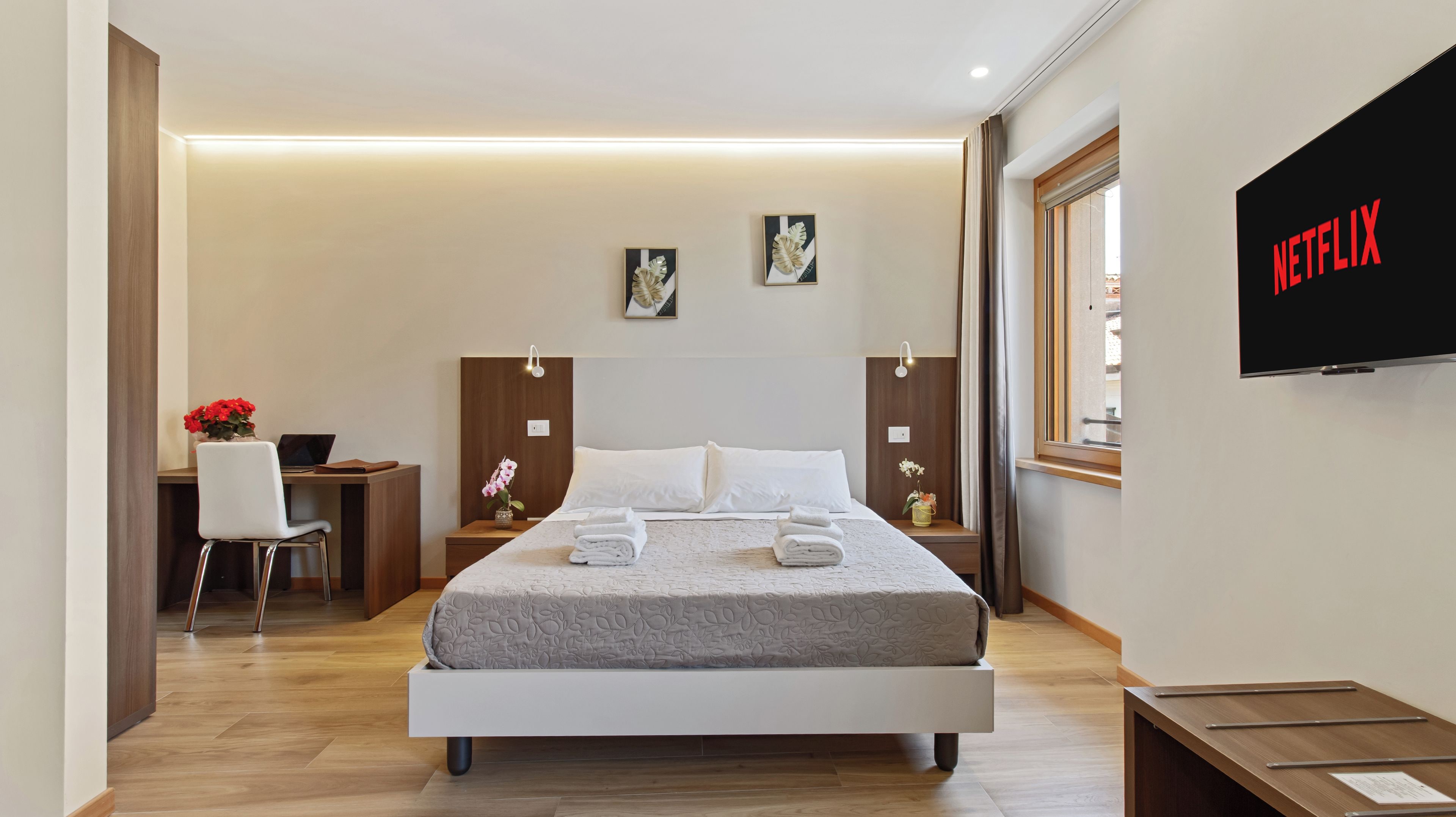 A modern hotel room with a bed, a desk, a chair, a TV, and artwork on the wall.