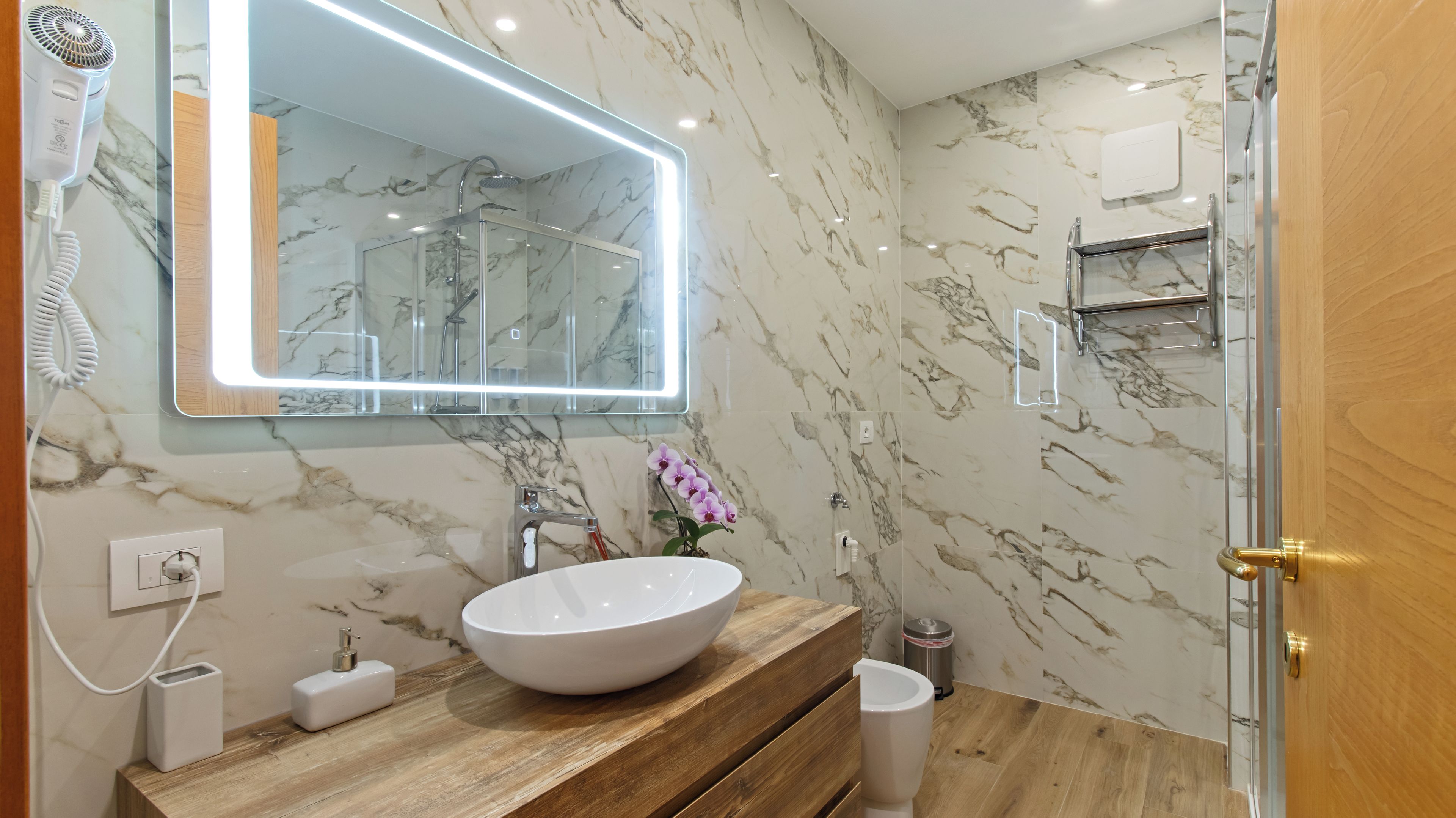 Ein Badezimmer mit Marmorwand, Waschtisch aus Holz, weißem Waschbecken, Duschkabine aus Glas und einem Spiegel mit integrierter Beleuchtung.
