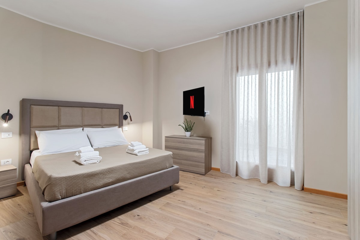 A modern hotel room with a bed, bedside tables, a TV, and a window with curtains.