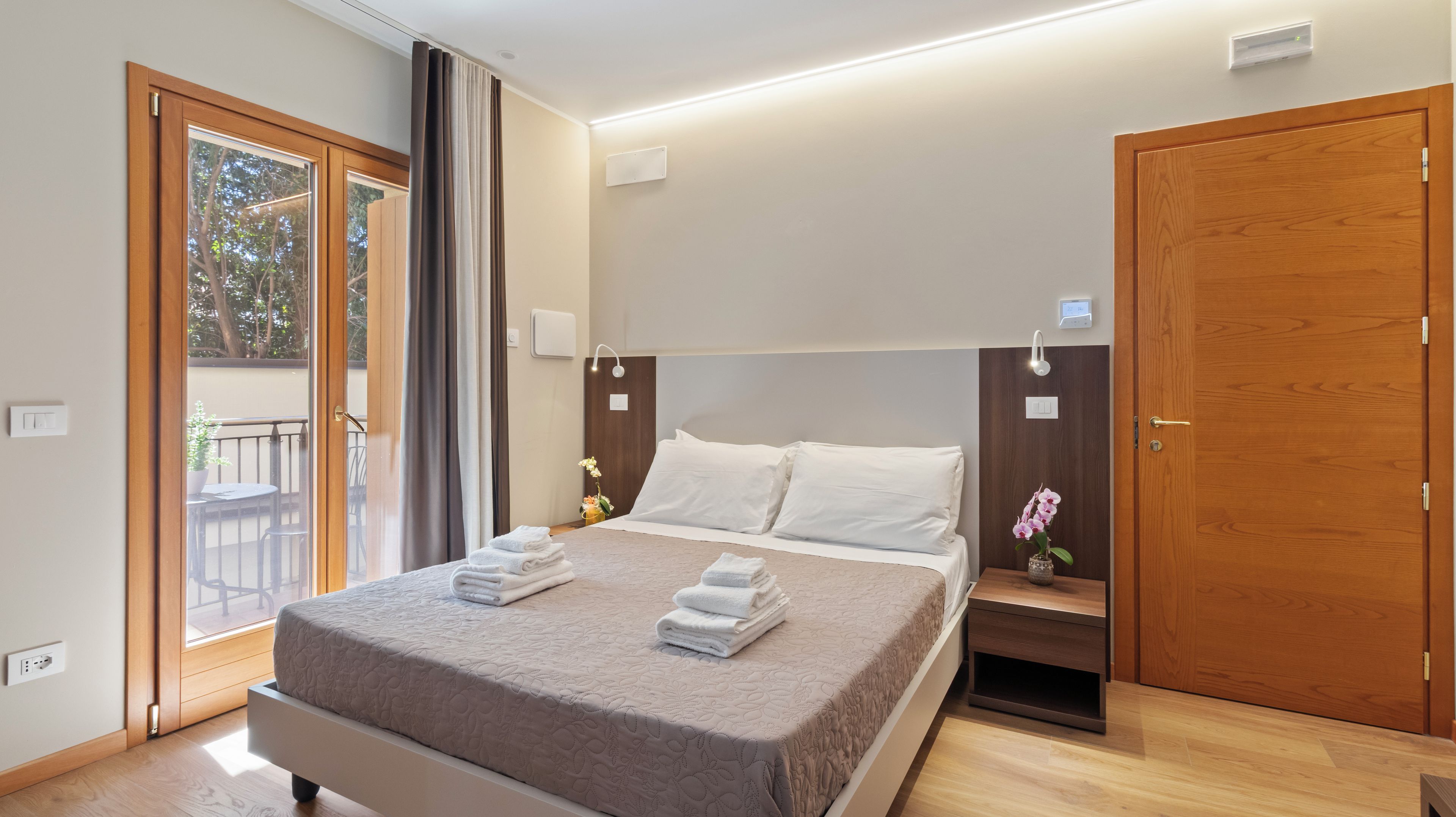 Una habitación de hotel moderna con una cama grande, un armario de madera, una mesita de noche con un jarrón de flores y un balcón con vistas a árboles.