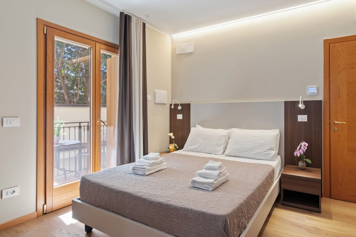 A modern hotel room with a large bed, a wooden wardrobe, a bedside table with a vase of flowers, and a balcony with a view of trees.