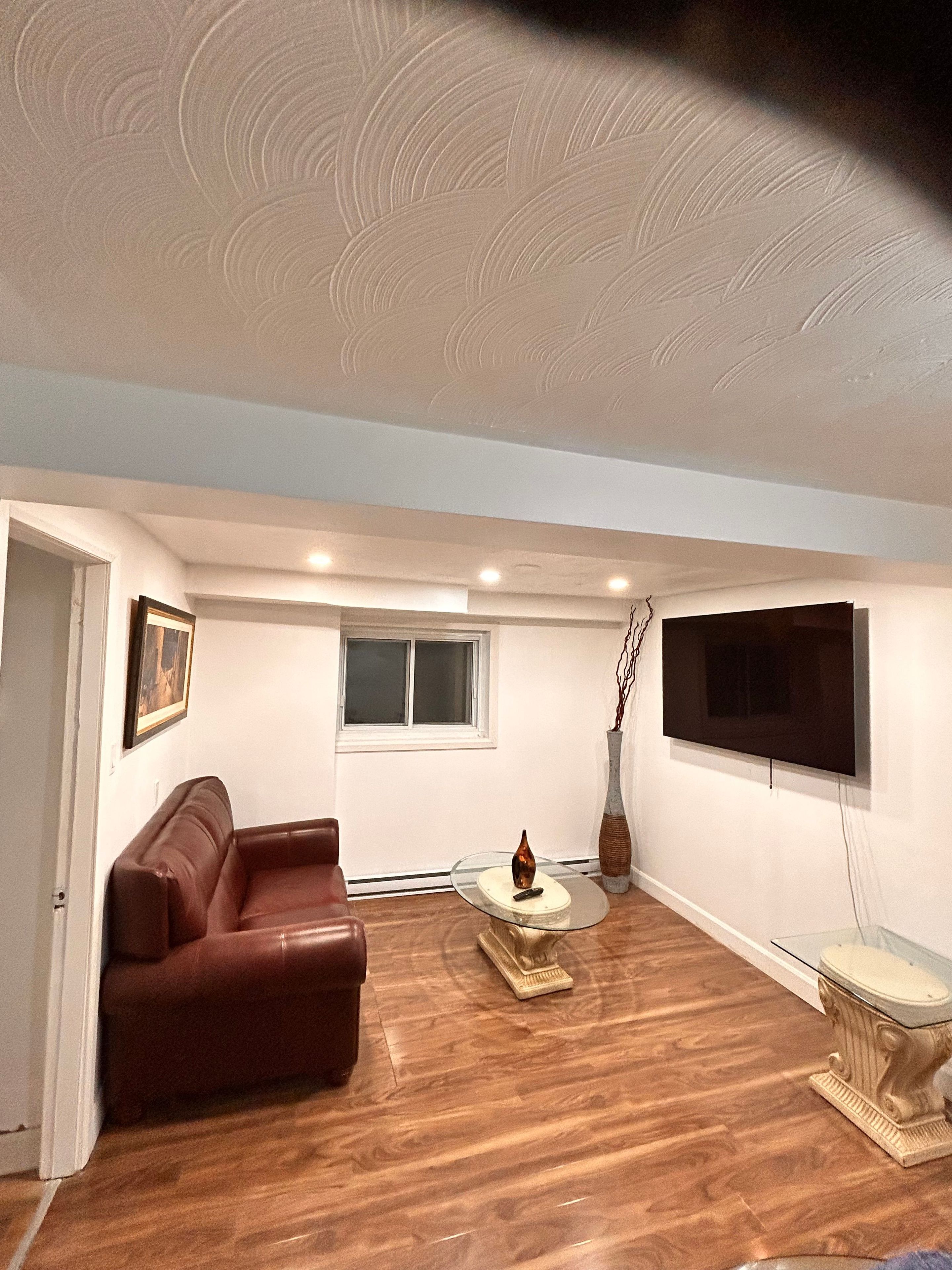 A living room with a brown leather couch, a glass coffee table, a flat-screen TV mounted on the wall, and a window with blinds.