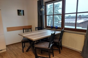 A dining area with a wooden table and chairs, a window with a view of buildings, a radiator, and a wall-mounted clock. - Small holiday home Allgäublick (Bad HIndelang)