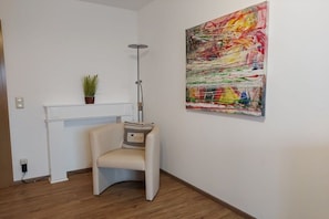 A room with a white desk, a chair, a floor lamp, a potted plant, and a colorful abstract painting on the wall. - Small holiday home Allgäublick (Bad HIndelang)