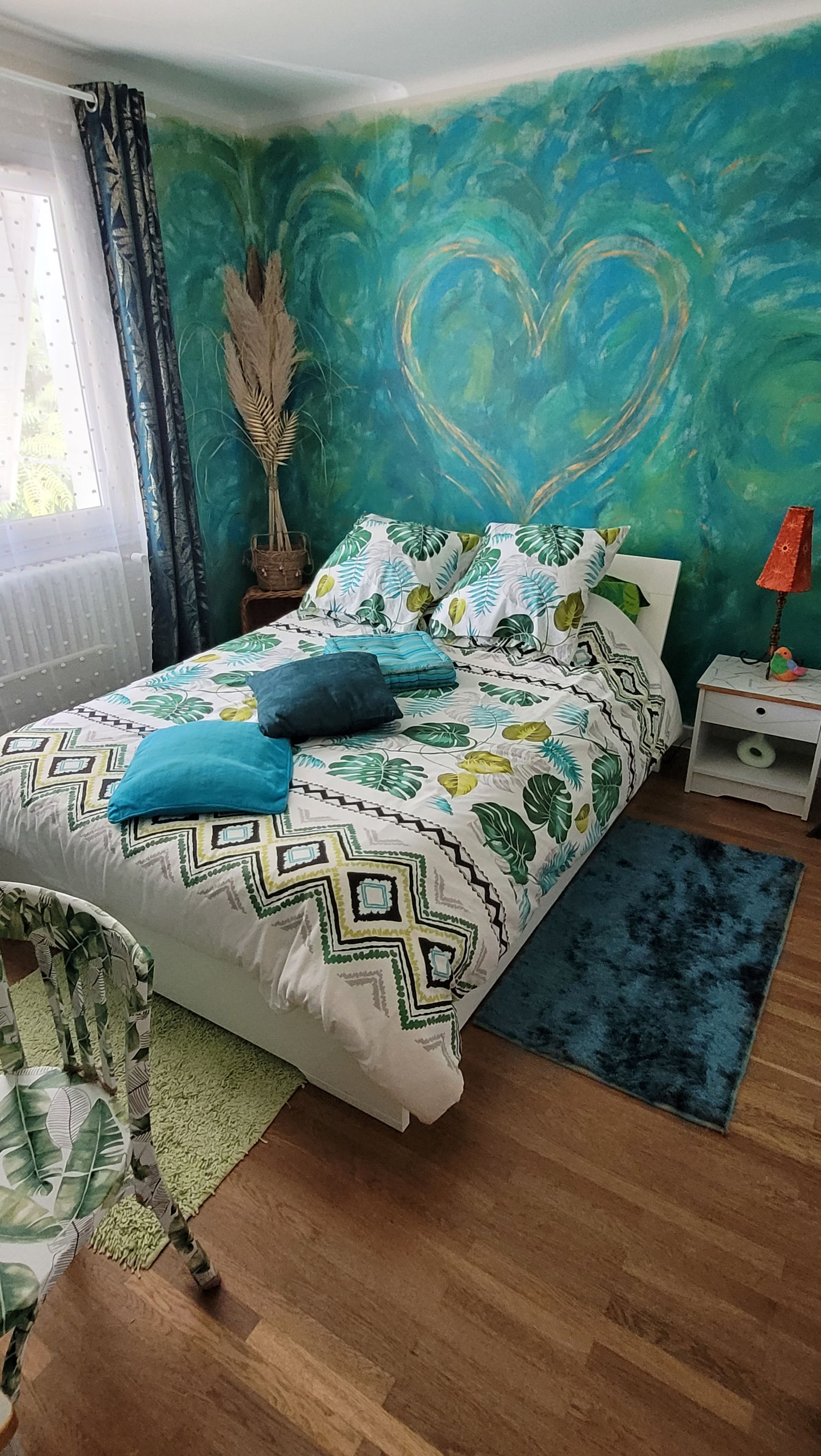 Une chambre à coucher avec un lit, une table de chevet, une lampe, une chaise et un mur décoré d’un motif en forme de cœur.
