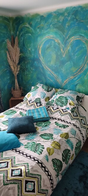 Ein Bett mit einem Bettbezug mit tropischem Blattmuster, einem herzförmigen Wandbild und einer Pflanze auf einem Nachttisch.