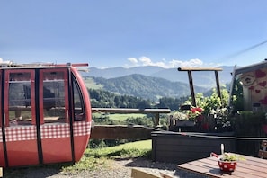 A red cable car with a checkered pattern, a wooden table with a red polka-dotted bowl, and a view of a mountainous landscape. - Oberstaufen with hotel use (Oberstaufen)