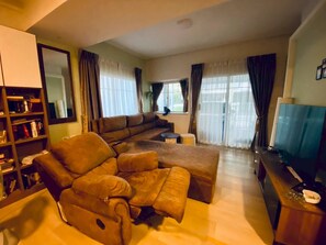 A living room with a sectional sofa, a television, a bookshelf, and a large window with curtains. - Serene 2-Bedroom House Rental in Bang Kaeo, Thailand (Tambon Bang Kaeo)