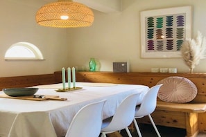 A dining area with a table set for four, a woven light fixture above, a framed botanical print on the wall, and a window with a semi-circular view.