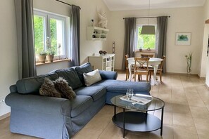 Ett vardagsrum med en hörnsoffa, ett runt soffbord, ett matbord med stolar och ett fönster med gardiner.