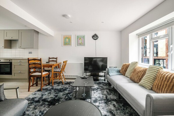 A modern living room with a sofa, a coffee table, and a TV. There is a dining area with chairs and a table. The room has a large window and a kitchen in the background. - Stylish Stay in Brighton London Road Gem (Brighton and Hove)