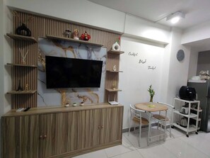 A modern living room with a flat-screen TV, wooden shelving, a small dining table, and a kitchen area with a coffee machine. - Fully furnished, modern Condo unit in ARC tower-Cebu City (Cebu City)