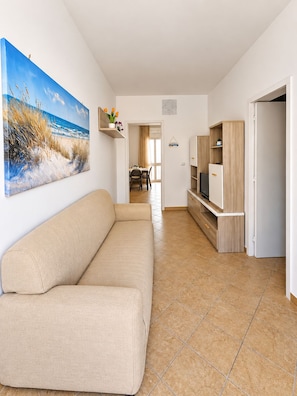 A living room with a beige sofa, a flat-screen TV, and a painting of a beach scene. - Casa Brezza – Private Garden Porto Cesareo (Porto Cesareo)
