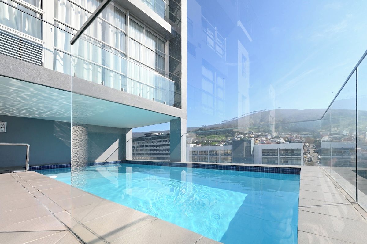 A rooftop swimming pool with clear blue water, surrounded by a glass barrier, and a view of a cityscape and hills in the background.