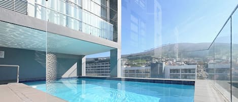 A rooftop swimming pool with clear blue water, surrounded by a glass barrier, and a view of a cityscape and hills in the background.