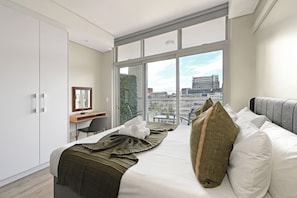 A modern hotel room with a large window, a bed with white linens, a desk, and a chair. - Cityscape Luxury Studio with Balcony, AC in Cape Town (Cape Town)
