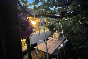 Taula de fusta amb cadires sota una pérgola, envoltada de verdura i un llum de llanternatge.