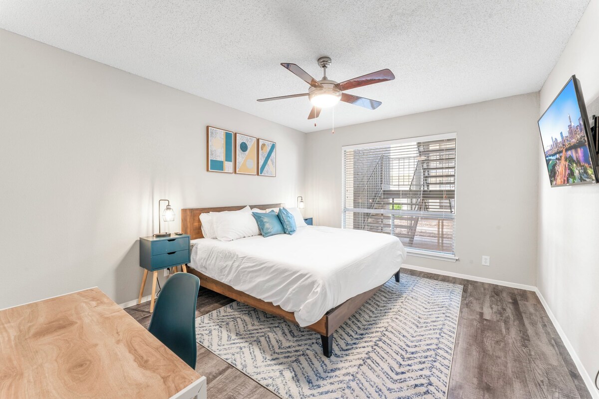A bedroom with a bed, a desk, a chair, a ceiling fan, and a window with blinds.