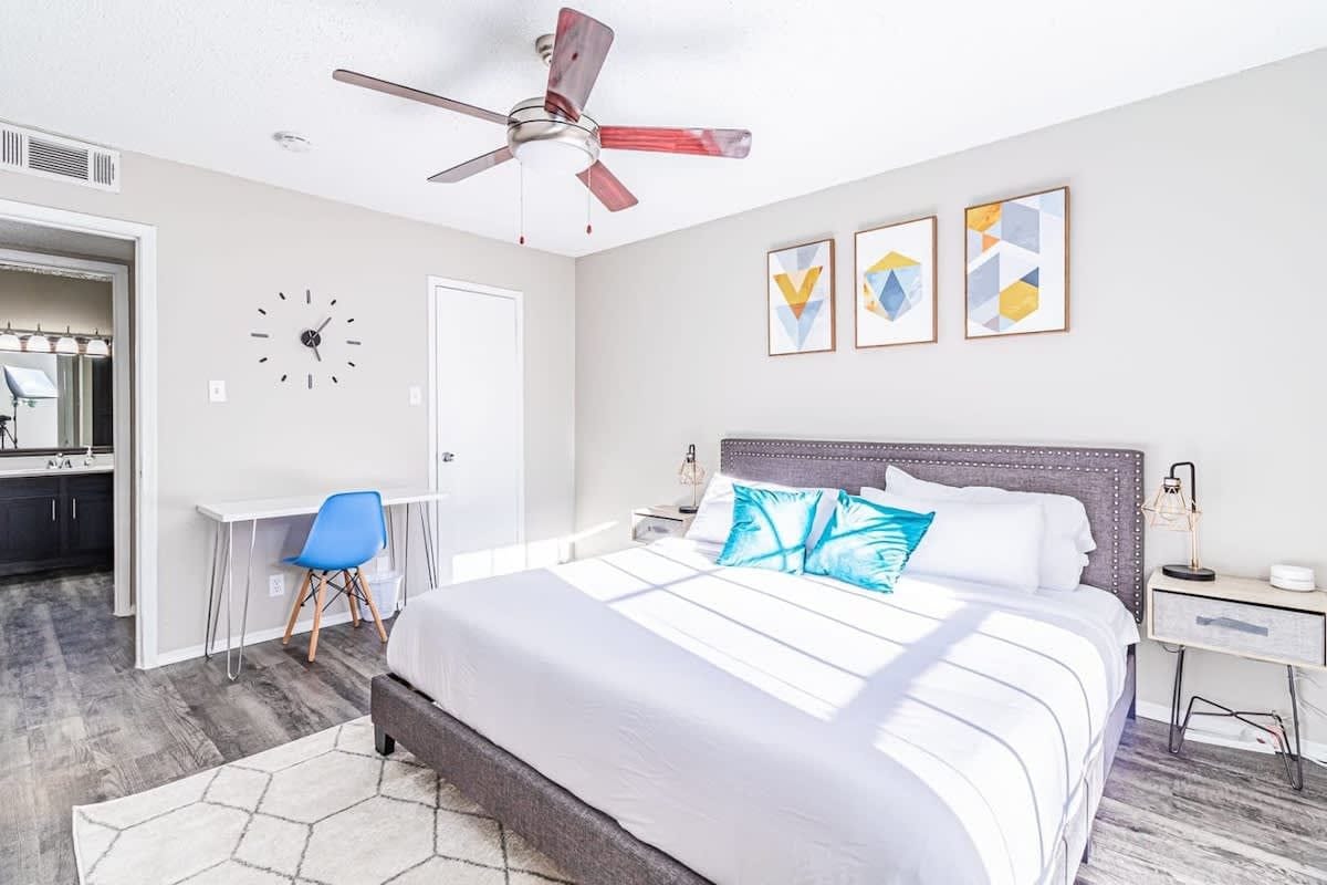 A bedroom with a bed, a desk, a chair, a ceiling fan, and artwork on the wall.