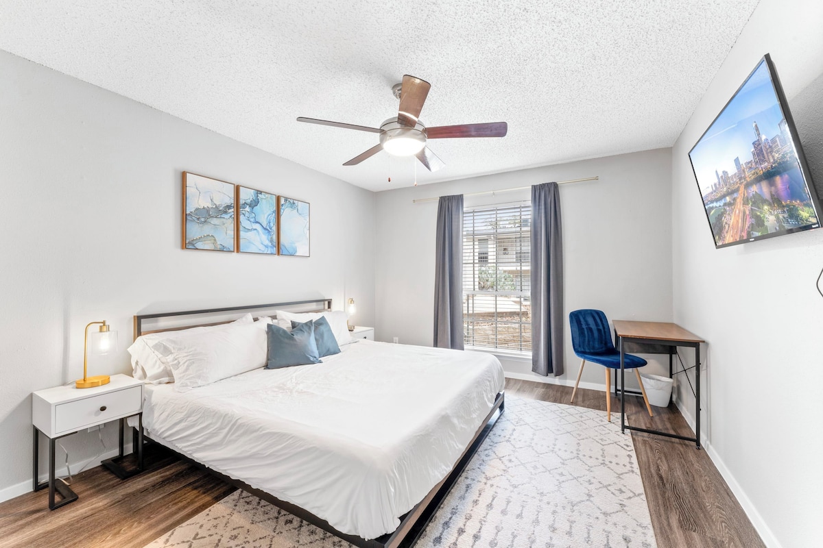 A modern bedroom with a bed, bedside tables, a desk, a chair, a ceiling fan, and a cityscape view.