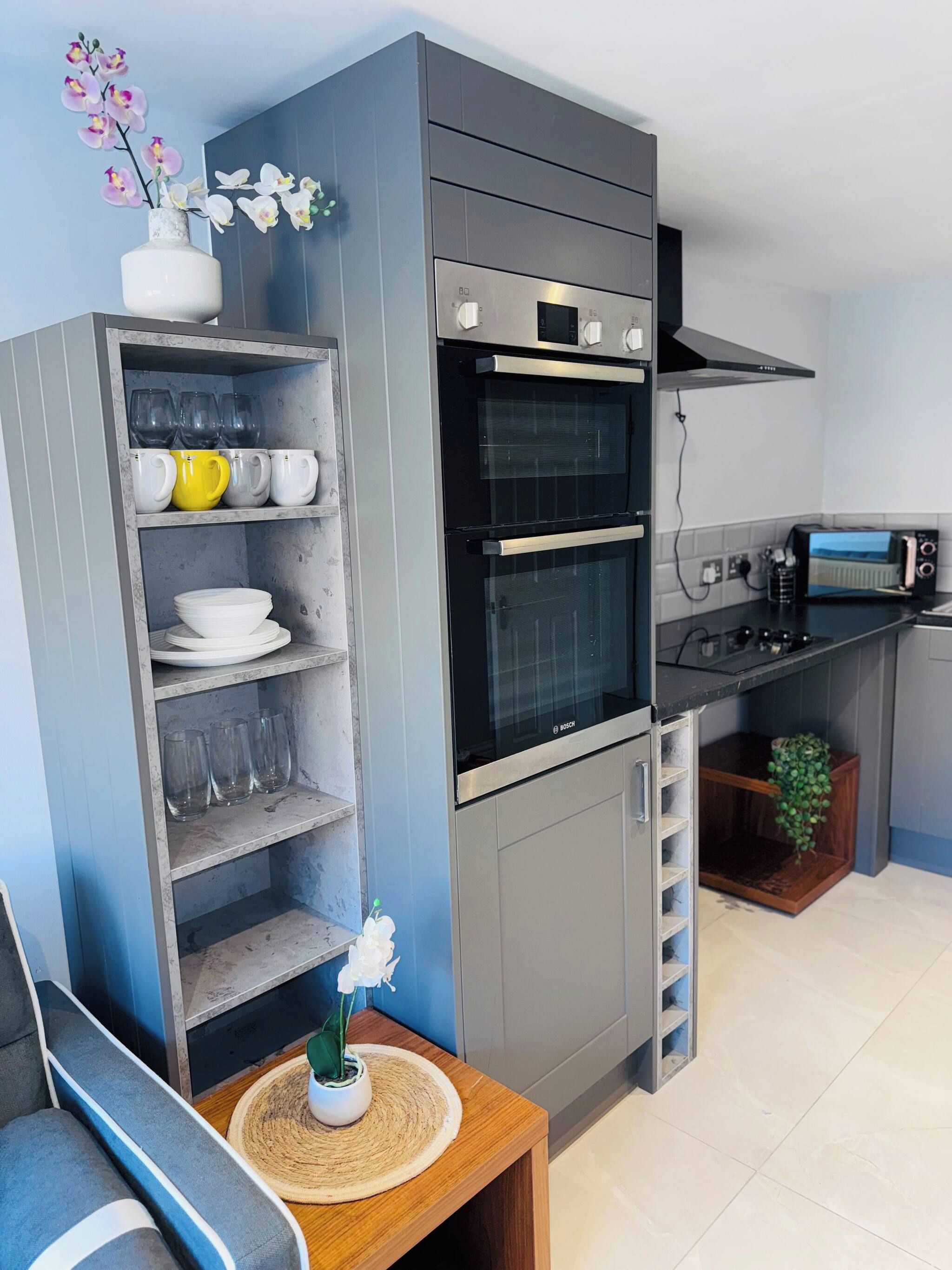 A compact kitchen with a built-in oven, a toaster, and a shelf unit with glassware and dishes.