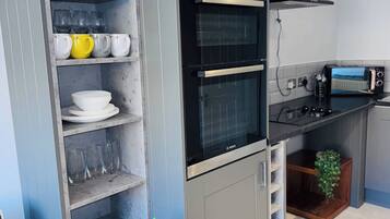 A compact kitchen with a built-in oven, a toaster, and a shelf unit with glassware and dishes.