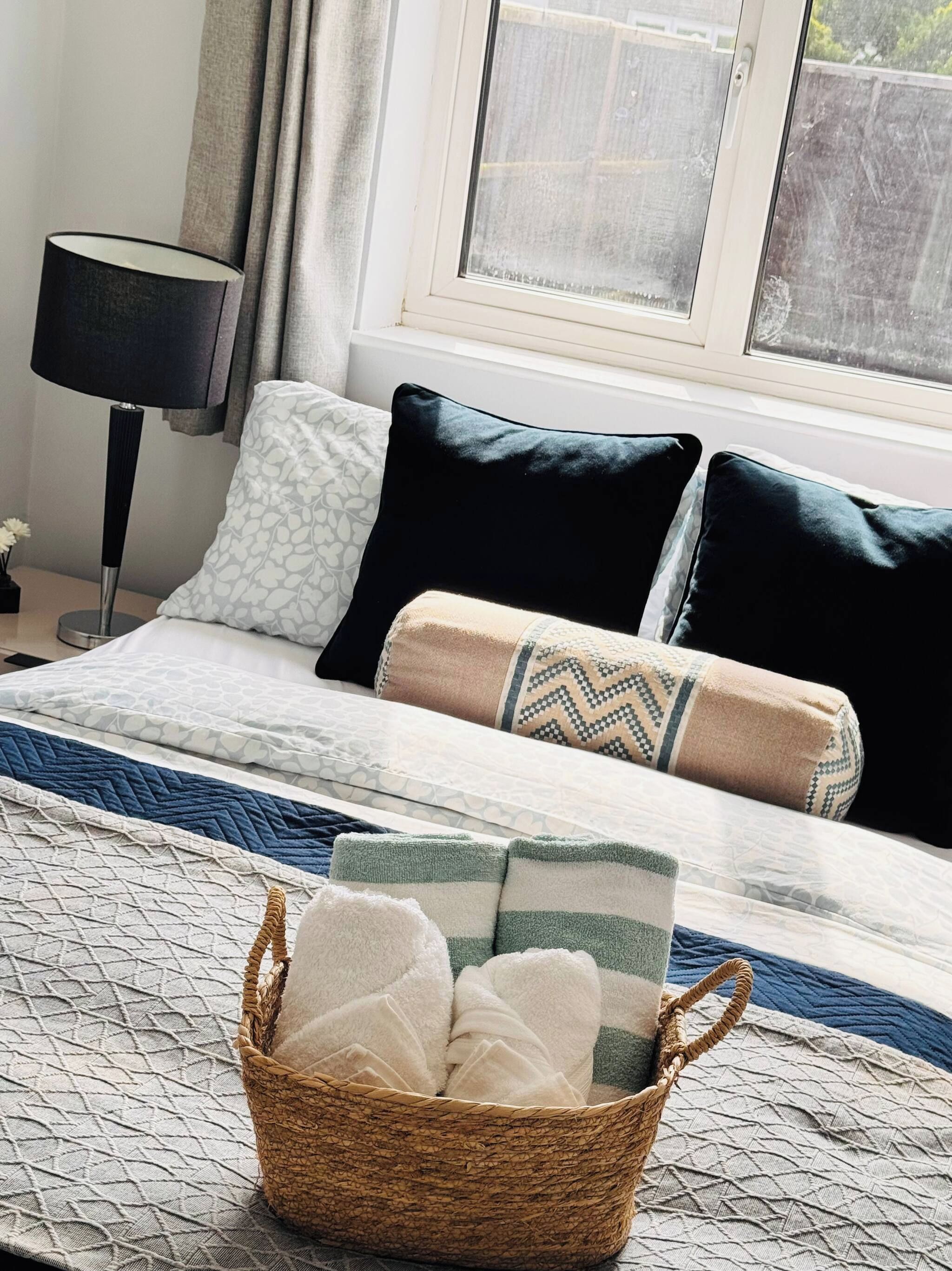 A neatly made bed with patterned bedding, a bedside lamp, and a basket of folded towels.