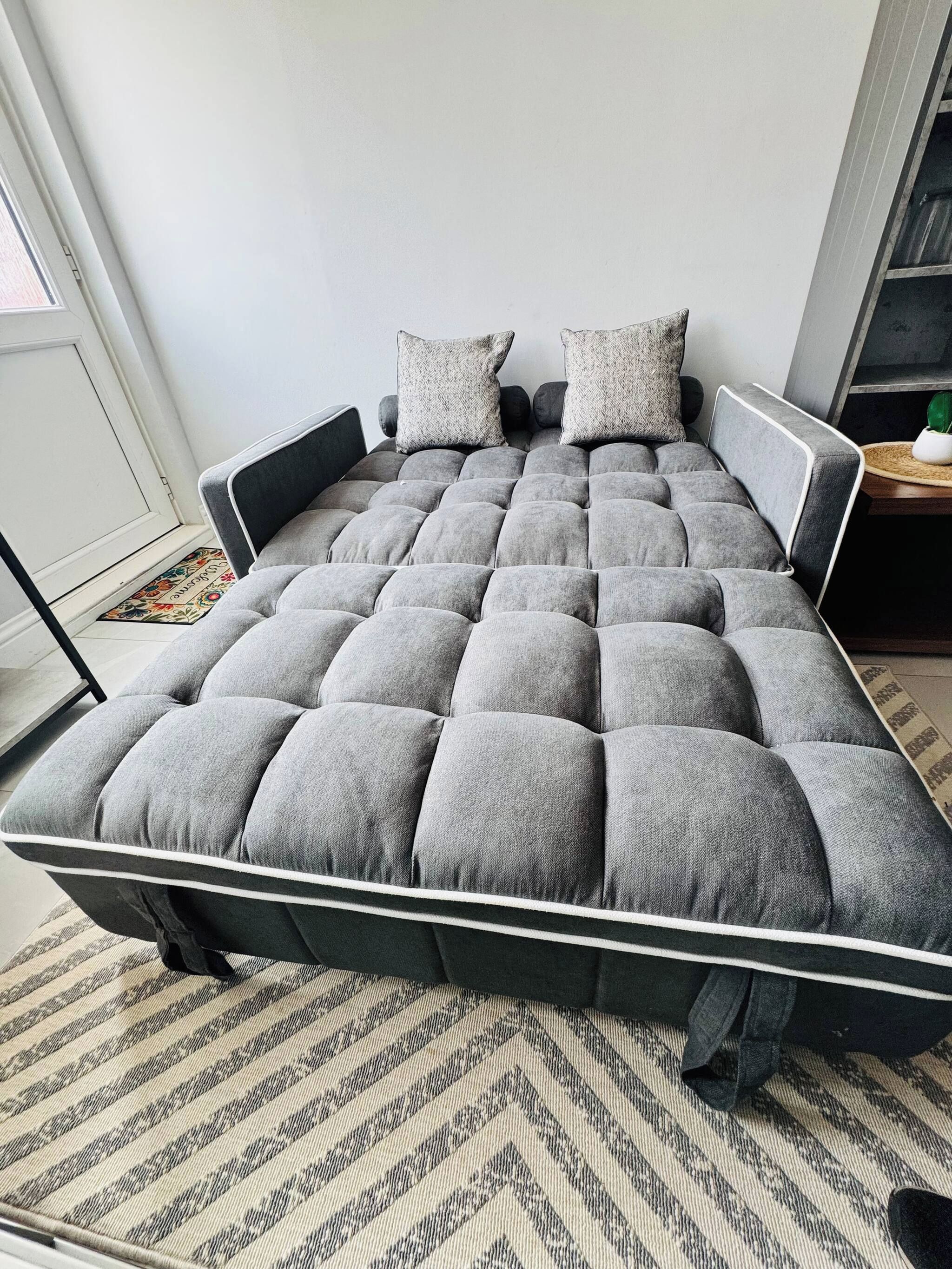Un lit avec une tête de lit et un pied de lit recouverts d’un tissu gris, deux oreillers et un tapis à rayures sur le sol.