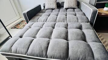 Un lit avec une tête de lit et un pied de lit recouverts d’un tissu gris, deux oreillers et un tapis à rayures sur le sol.