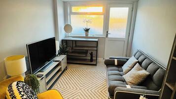 A modern living room with a sofa, a television, a small table with a lamp, and a window with a view.