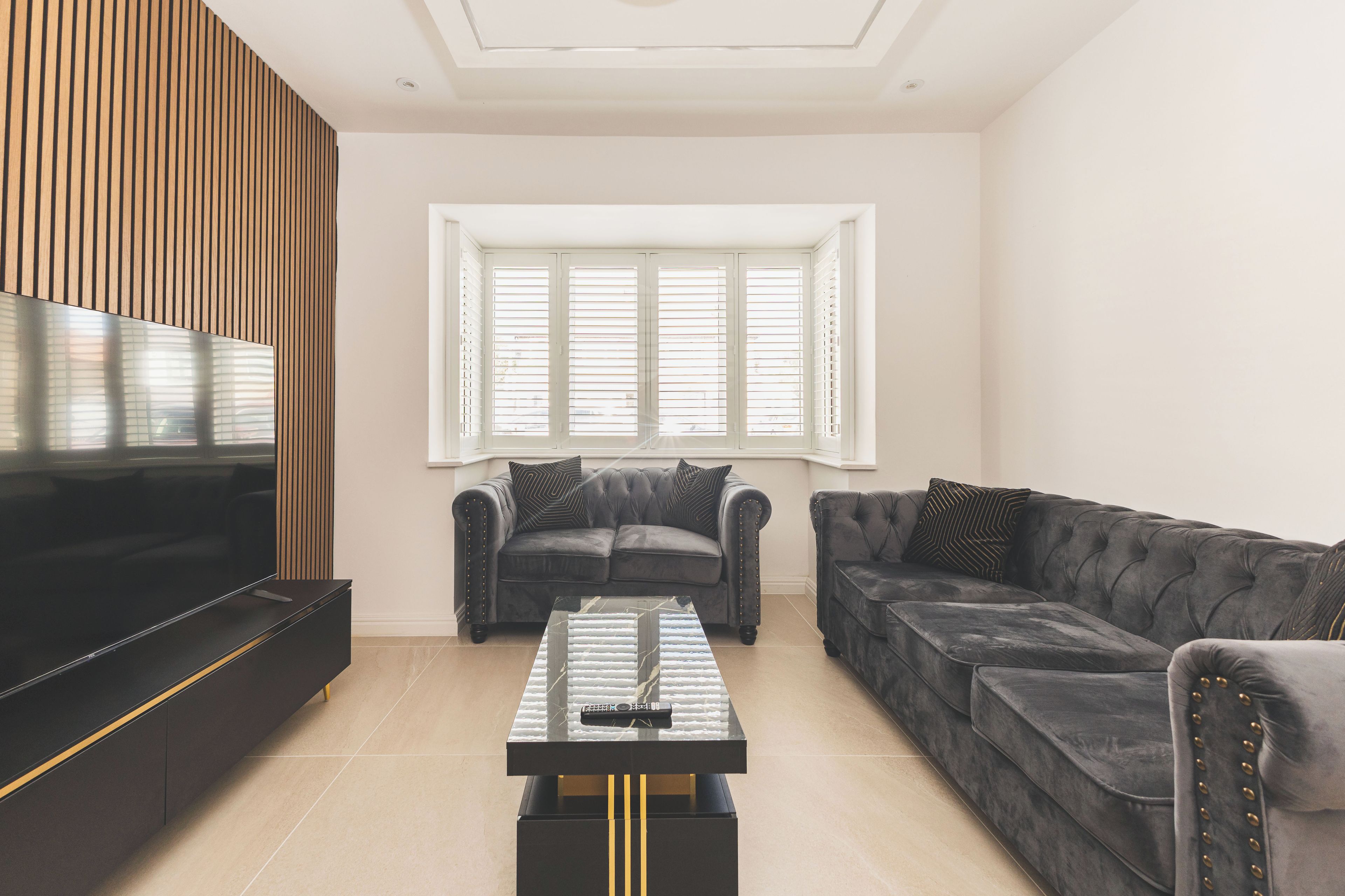 A modern living room with a flat-screen TV, two sofas, a coffee table, and a large window with blinds.