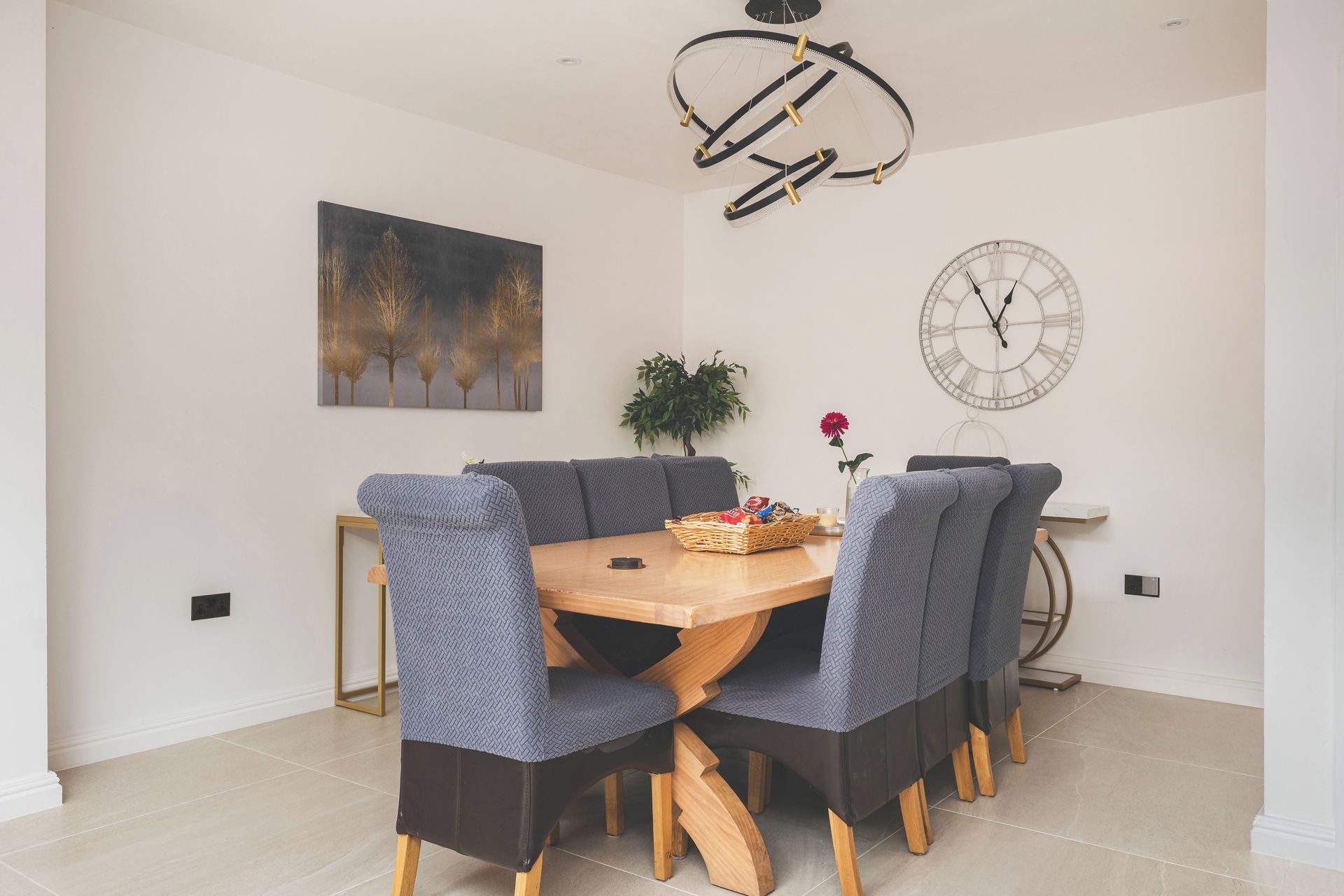 A dining area with a round wooden table, grey upholstered chairs, a wall-mounted clock, and a painting of trees.