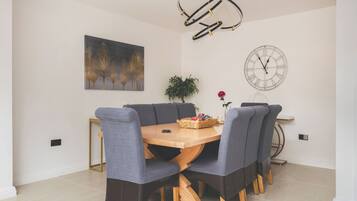 A dining area with a round wooden table, grey upholstered chairs, a wall-mounted clock, and a painting of trees.