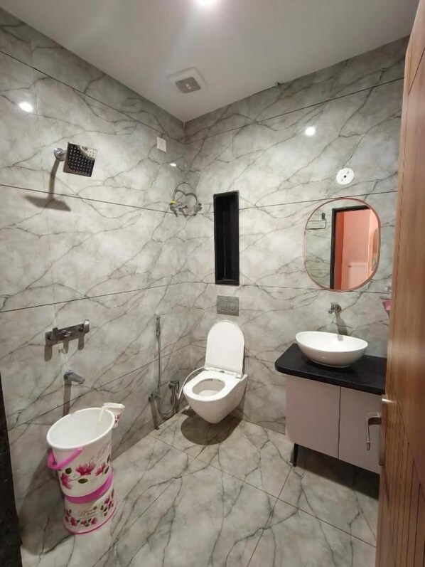 A bathroom with a toilet, sink, mirror, shower, and a decorative bucket. - Hotel Kanak Shree (Omkareshwar)