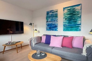 Ein modernes Wohnzimmer mit einem grauen Sofa, bunten Kissen, einem Flachbildfernseher und abstrakten Bildern an der Wand.