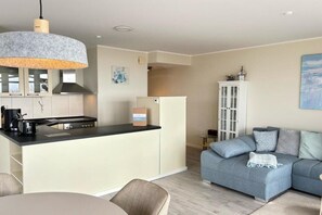 A modern kitchen with a central island, a dining area with a round table, and a living room with a blue sofa. - Holiday apartment with 1 bedroom (Großenbrode)