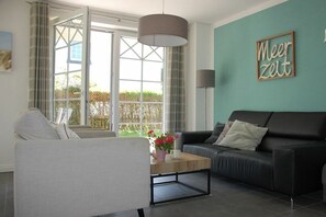 Ein modernes Wohnzimmer mit Sofa, Sessel und Couchtisch. Ein großes Fenster mit Vorhängen, eine Pendelleuchte und ein an der Wand befestigtes Schild.