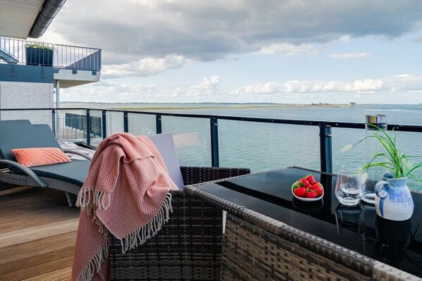 Un balcon avec vue sur la mer, une chaise en osier, une table avec un saladier de fraises et un vase avec une plante.