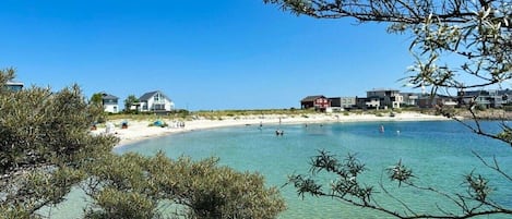 A beach with clear water, a sandy shore, and buildings in the background.