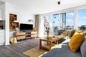 Un salon moderne doté d’un téléviseur à écran plat, d’étagères en bois, d’un canapé avec des coussins, d’une table basse et d’une grande fenêtre offrant une vue sur la ville.